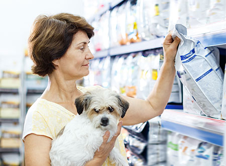 woman viewing pet food bag on shelf while holding dog in arm